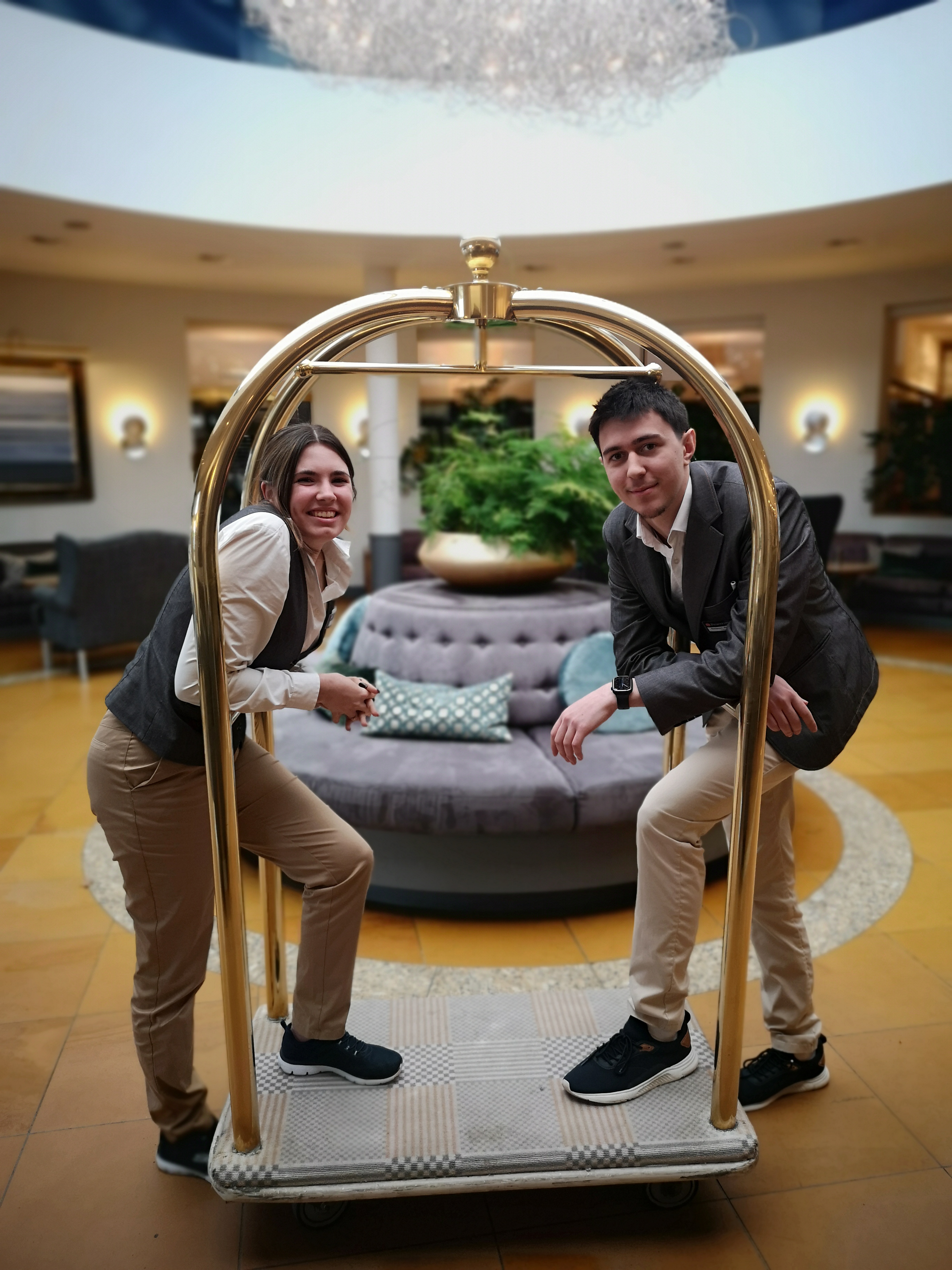zwei Auszubildende auf Gepäckwagen, der in der Hotelhalle steht.